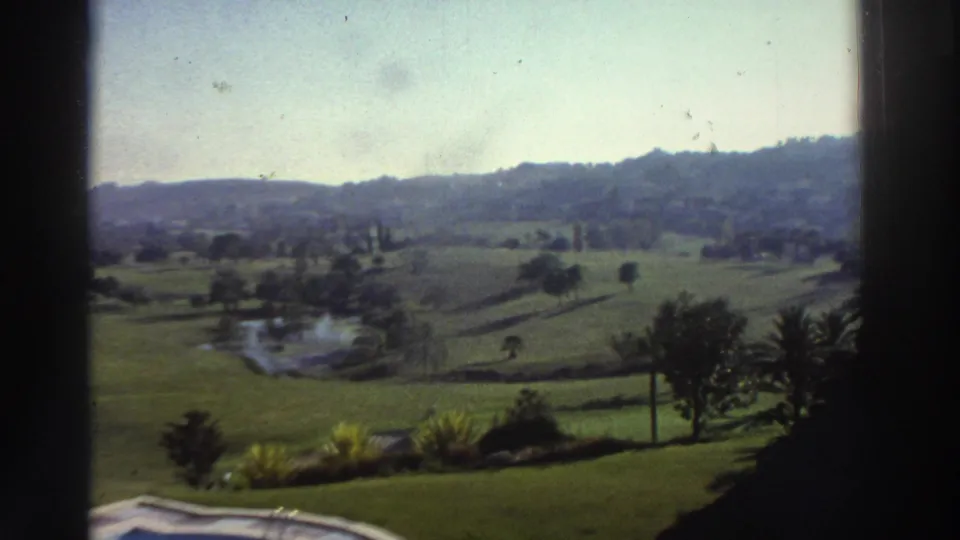 1976 Greece 8mm frame showing a mostly static view of landscaped grounds, a pond, trees, and hills, with the pool partly visible below.