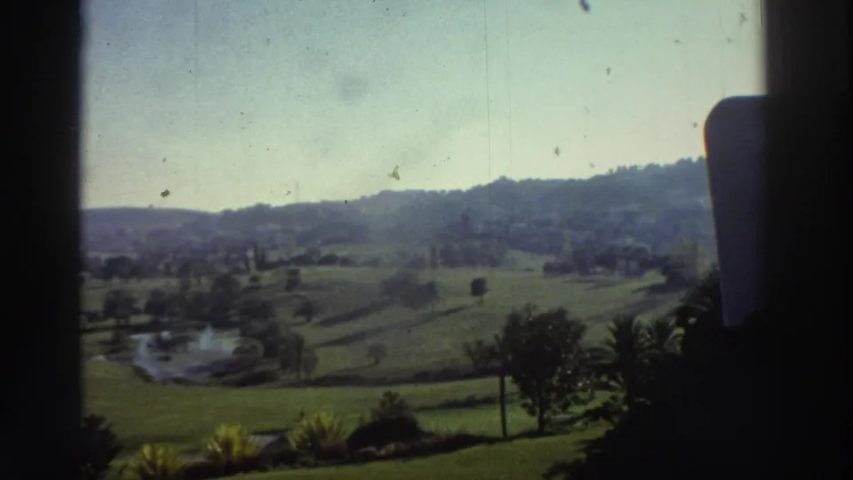 1976 Greece home-movie frame looking past a dark interior edge toward lawns, a small pond, trees, and distant hills under bright sky.