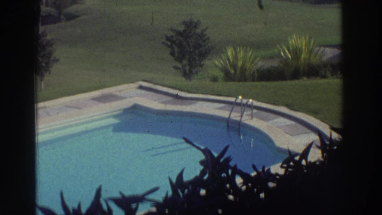 1976 Greece 8mm frame showing a quiet pool corner with clear blue water, pale deck, lawn, shrubs, and foreground foliage.