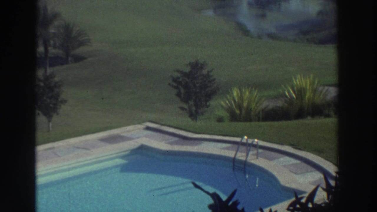1976 Greece 8mm frame with a shaped pool edge, metal ladder, lawn, small trees, and ornamental planting beyond the deck.