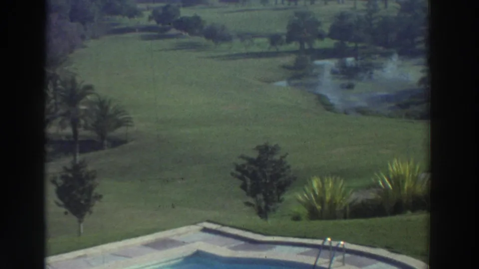 1976 Greece 8mm frame looking past the pool and garden plantings toward a green lawn, palm, trees, and distant water.