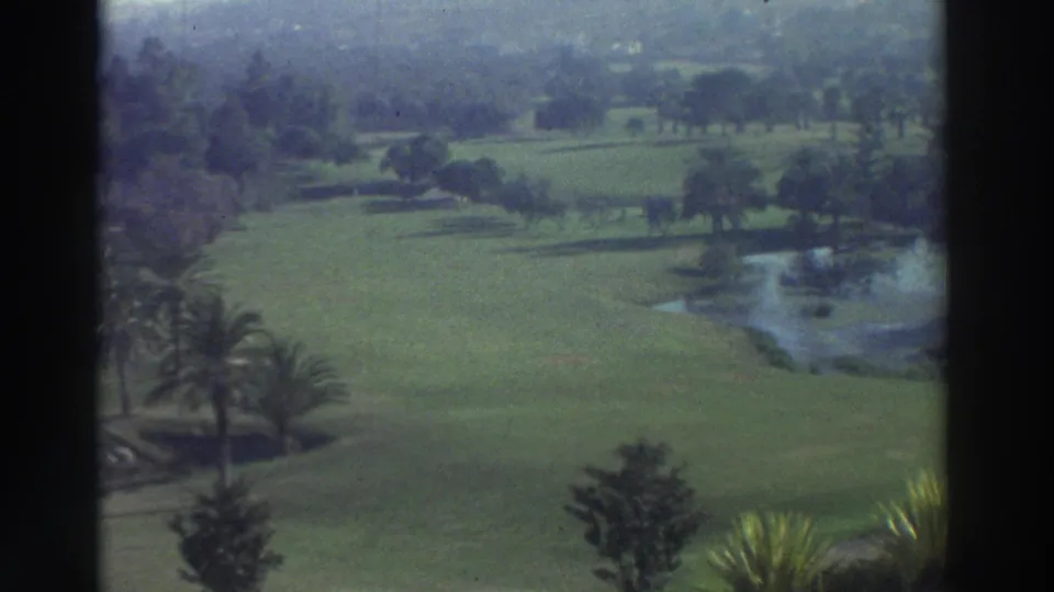 1976 Greece 8mm frame showing landscaped green grounds with a palm, scattered trees, reflective pond, and partial pool edge.