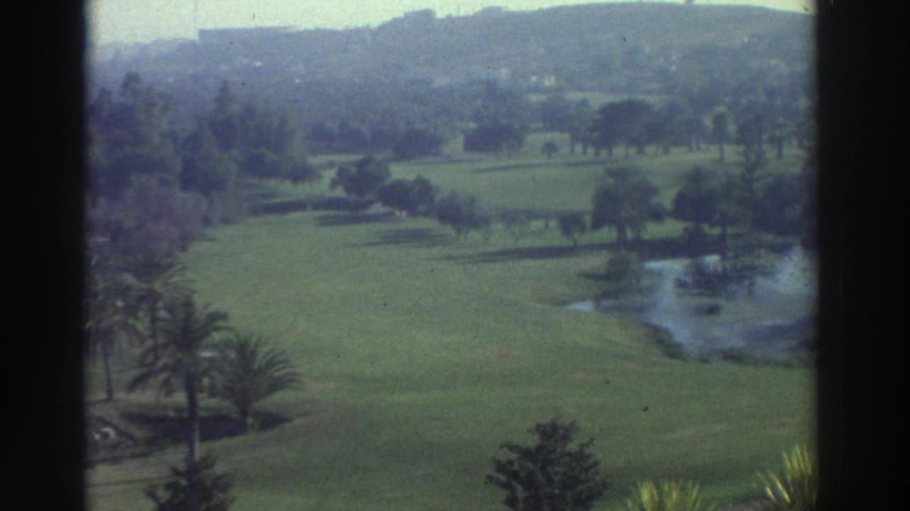 1976 Greece 8mm frame showing manicured green grounds, a pond, scattered trees, palms, and hazy hills in the distance.