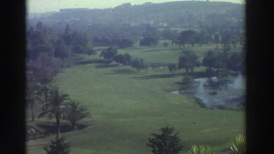 1976 Greece 8mm frame showing manicured green grounds, a pond, scattered trees, palms, and hazy hills in the distance.