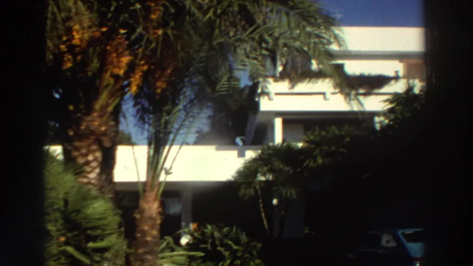 1976 Greece 8mm frame of a white multi-level villa behind palms and dense vegetation, with part of a parked car at the edge.