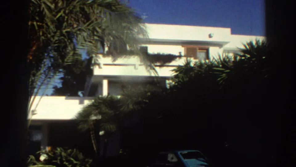 1976 Greece 8mm frame showing the villa facade, balconies or terraces, palm fronds, heavy shadows, and a parked car near the lower edge.