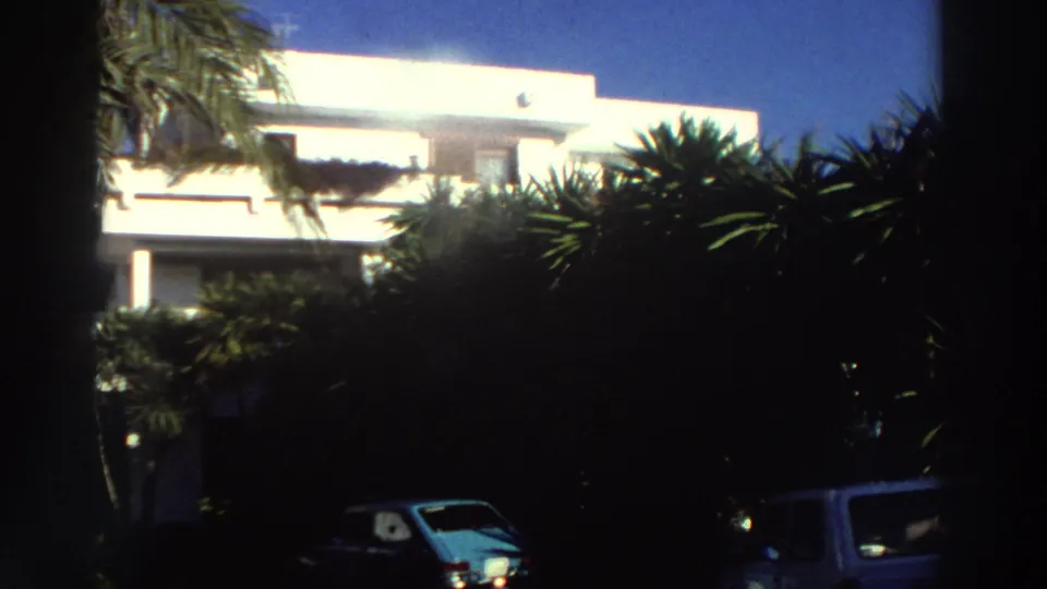 1976 Greece 8mm frame of the white villa partly hidden by palms and dark vegetation, with two parked cars visible near the foreground.
