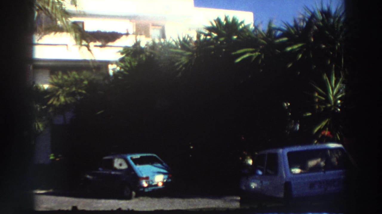 1976 Greece home-movie frame showing a white resort building, palms, parked cars, and dark foreground film edges.