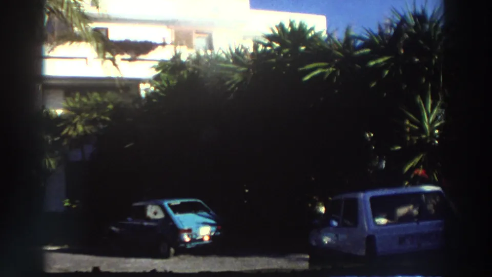 1976 Greece home-movie frame showing a white resort building, palms, parked cars, and dark foreground film edges.