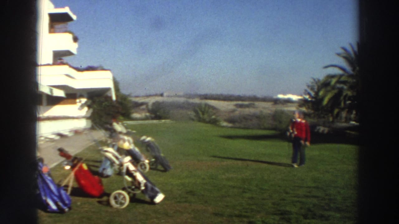 1976 Greece resort lawn with a woman in a red top walking past golf bags, carts, palm trees, and a white hotel.