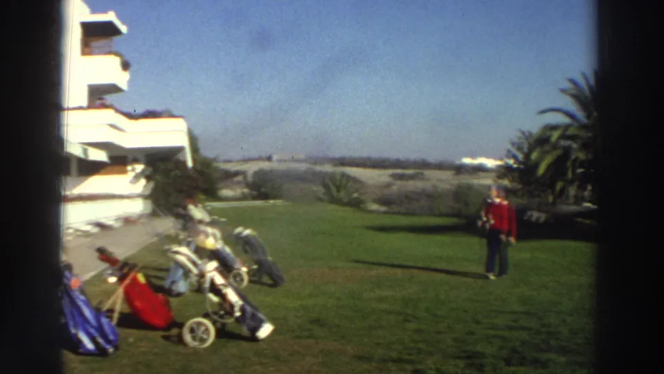 1976 Greece resort lawn with a woman in a red top walking past golf bags, carts, palm trees, and a white hotel.