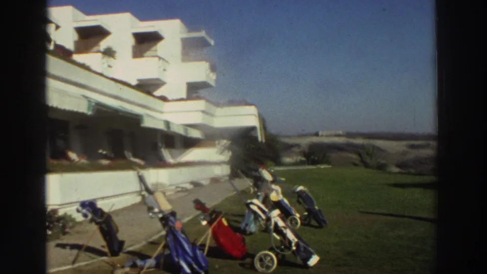 1976 Greece home-movie view of golf bags and carts arranged on a resort lawn beside a white hotel terrace.