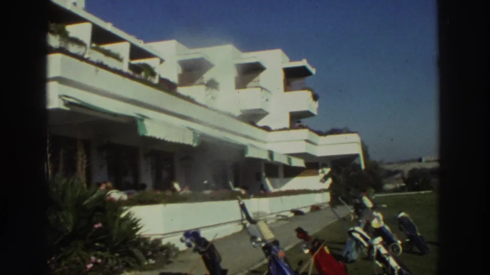1976 Greece resort frame showing white hotel balconies, a terrace, palm planting, and golf bags on the lawn.