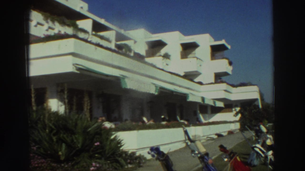 White modernist building with balconies, plants, scooters, and blue sky.