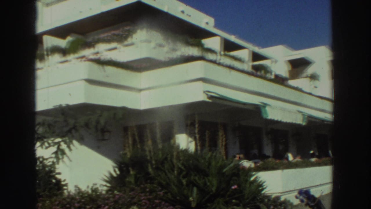 White balcony building with planters, awning, shrubs, and blue sky.