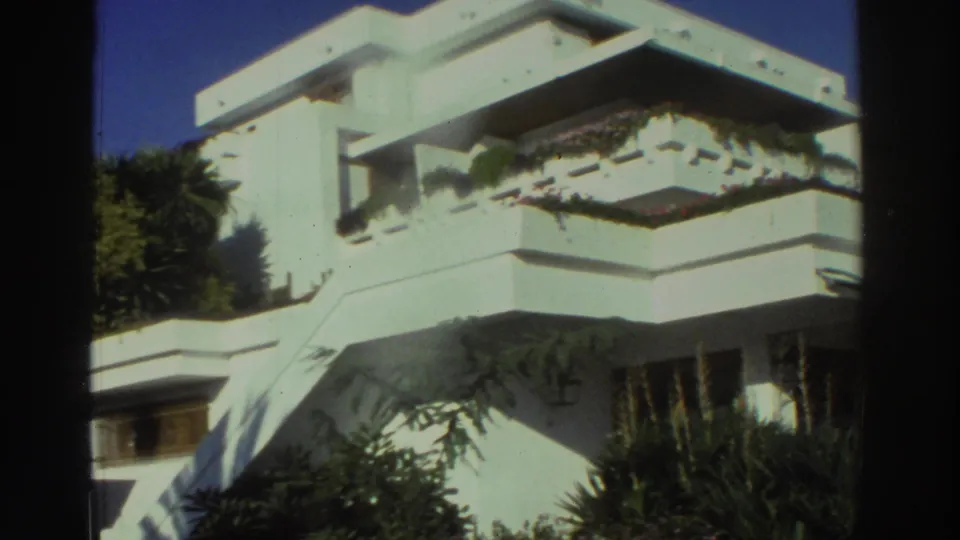 1976 Greece 8mm frame showing a closer exterior angle on white geometric balconies, planted terrace edges, trees, deep shadows, and foreground shrubs.