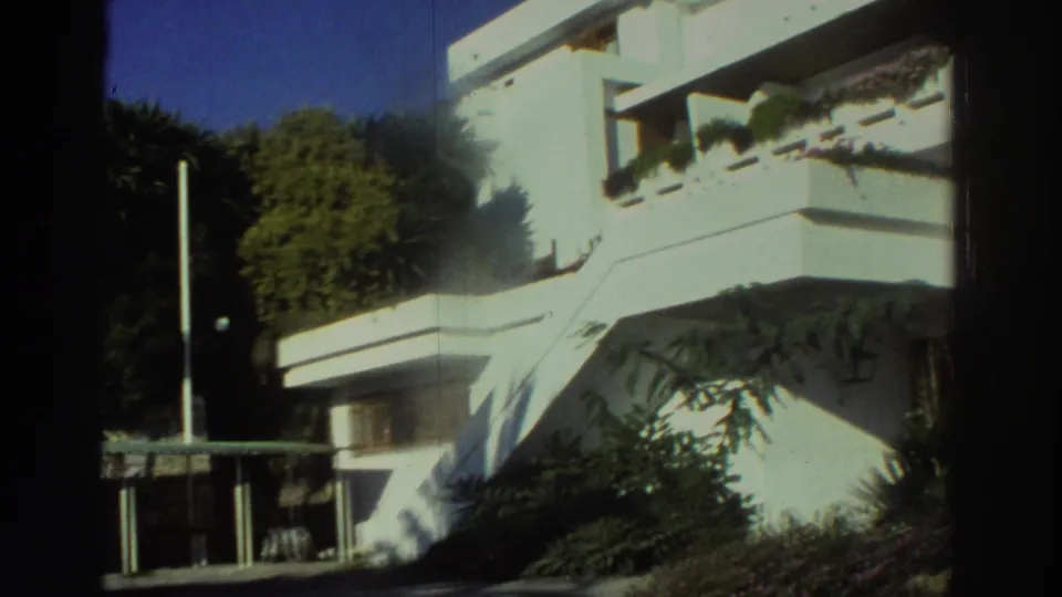 1976 Greece exterior frame with a white stairway, angular balconies, planted ledges, shrubs, trees, and a vertical pole in front of the building.