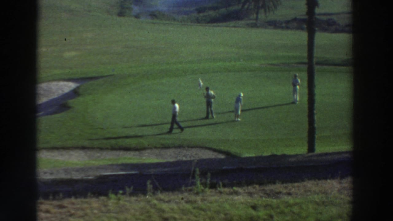 1976 Greece home-movie frame showing men on a sunlit golf course, with palm trunks, long shadows, and dark window-like edges.