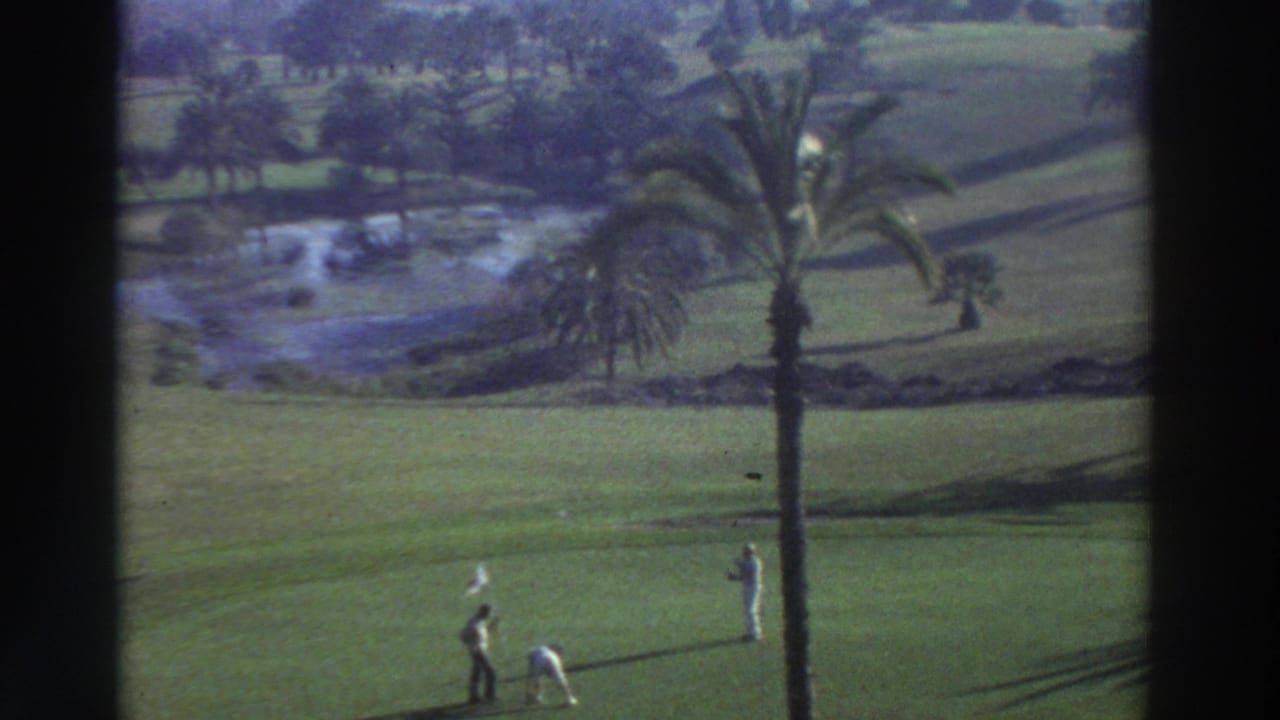 Wide 1976 Greece golf course view with players on the green, a palm tree, water hazard, and visible Super 8 grain.