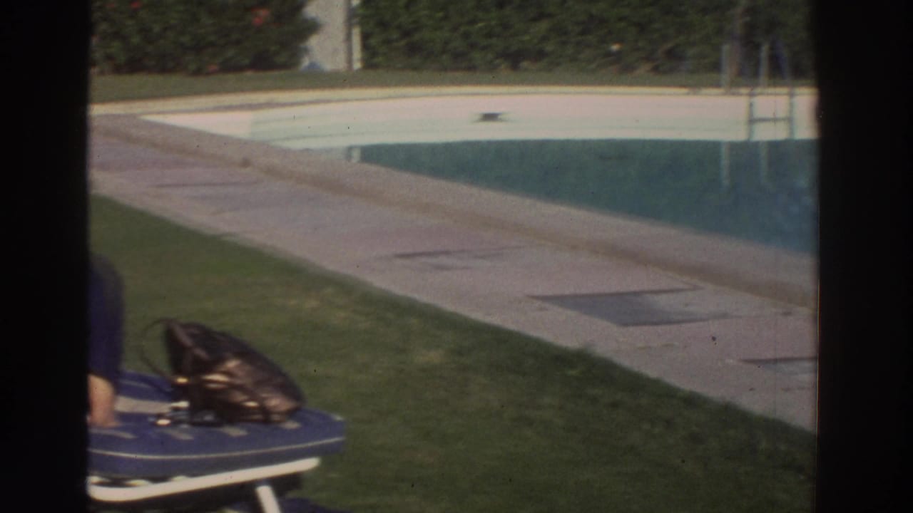 1976 Greece 8mm frame panning past the pool toward a lawn, lounge chair, handbag, and cropped adult figure.