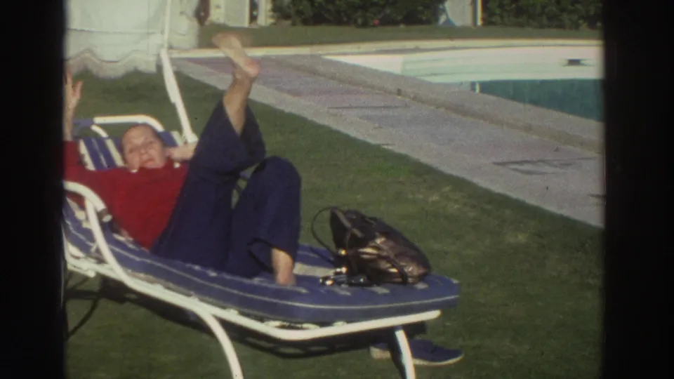 1976 Greece 8mm frame showing an adult woman reclining on a blue lounge chair near the swimming pool and waving toward the camera.