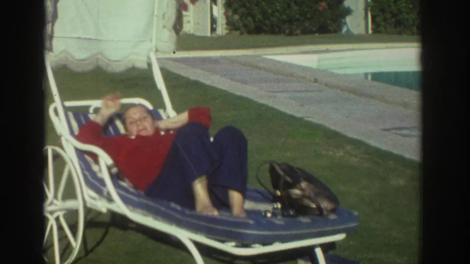 1976 Greece 8mm frame with an adult woman smiling and waving from a blue lounge chair near the swimming pool and lawn.