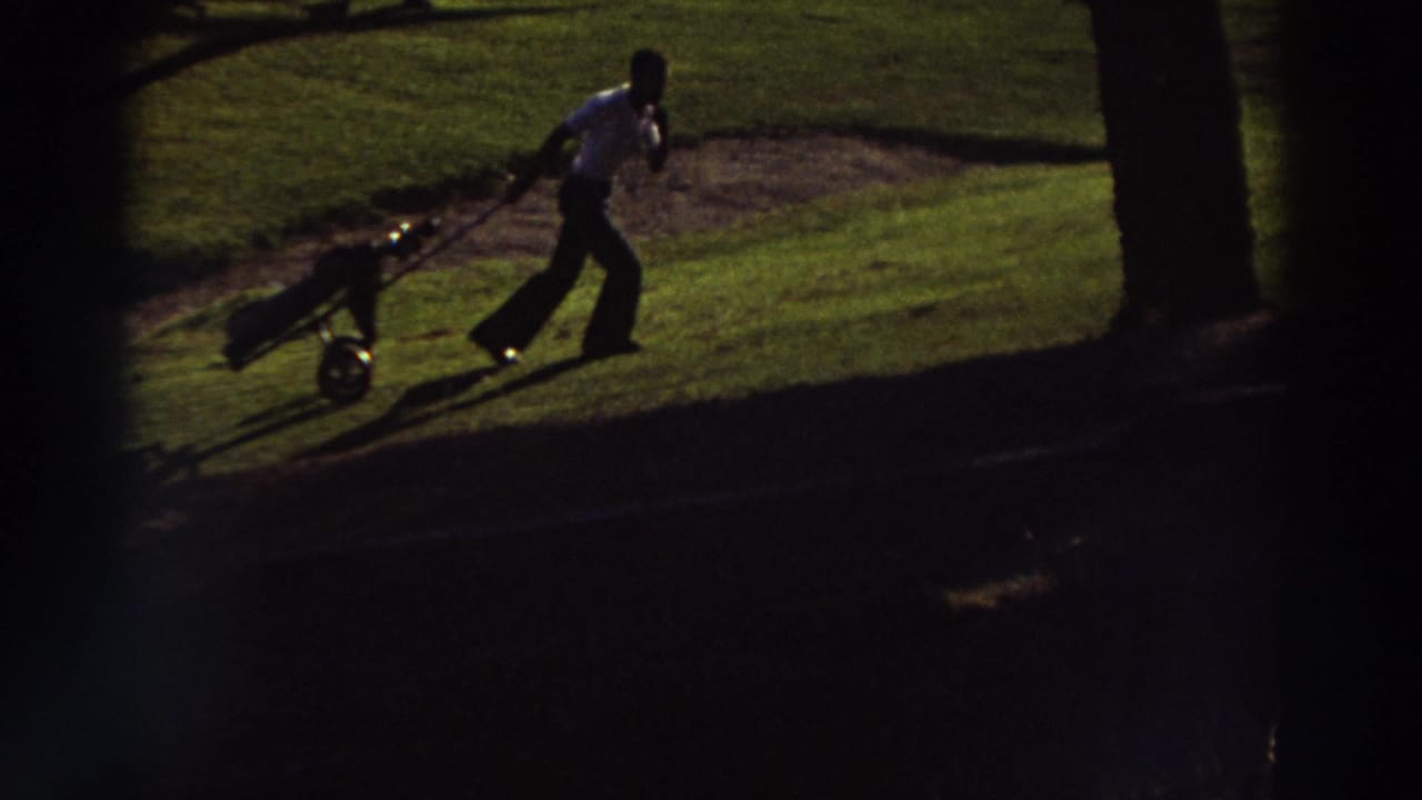 1976 Greece home-movie frame showing an adult man pulling a golf bag cart across a sunlit golf course with deep shadow and dark vignetting.