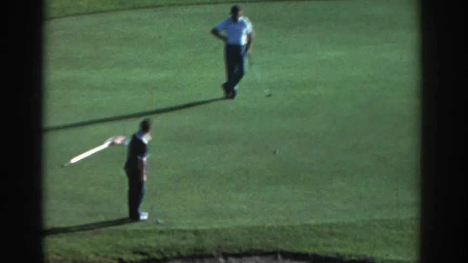 1976 Greece home-movie still showing two adult men on a golf green, with one holding a club and the other standing near the putting line.