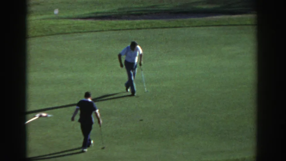 1976 Greece 8mm frame showing two adult men walking across the green after a putt, with long shadows and a sand bunker behind them.
