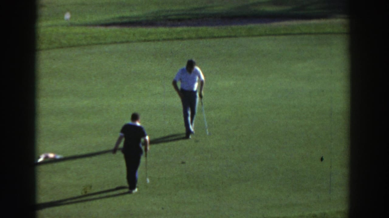 In 1976 Greece metadata, two adult men appear on a sunlit golf green, one preparing near the hole while another watches from behind.