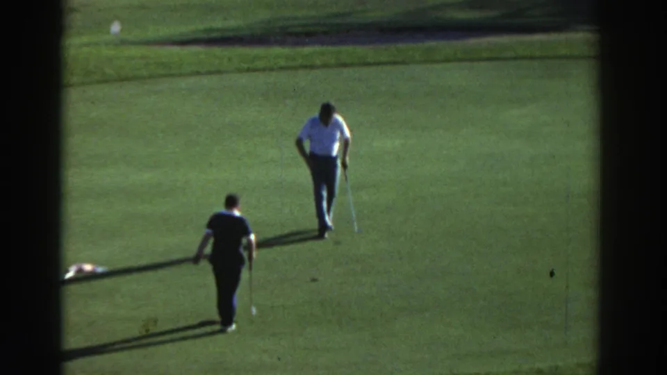 In 1976 Greece metadata, two adult men appear on a sunlit golf green, one preparing near the hole while another watches from behind.