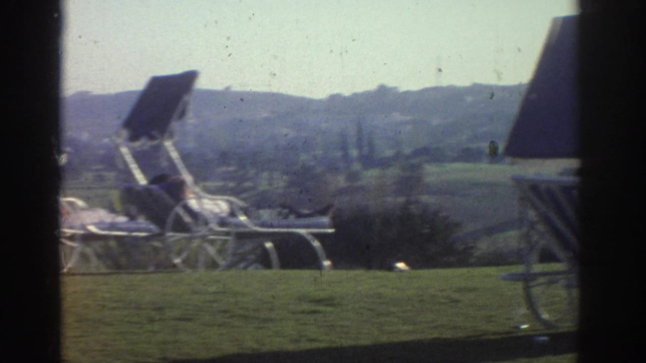 The camera frames wheeled lounge chairs and raised shades near shrubs, with a landscaped lawn and hazy hillside beyond.