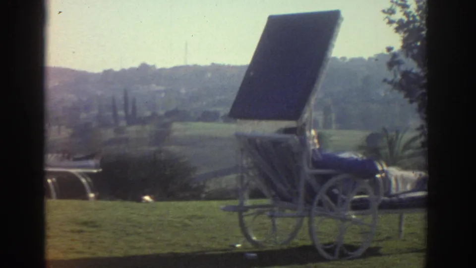 A wheeled outdoor lounge chair with a raised sun shade sits on a grassy area, backed by shrubs and a hazy hillside landscape.