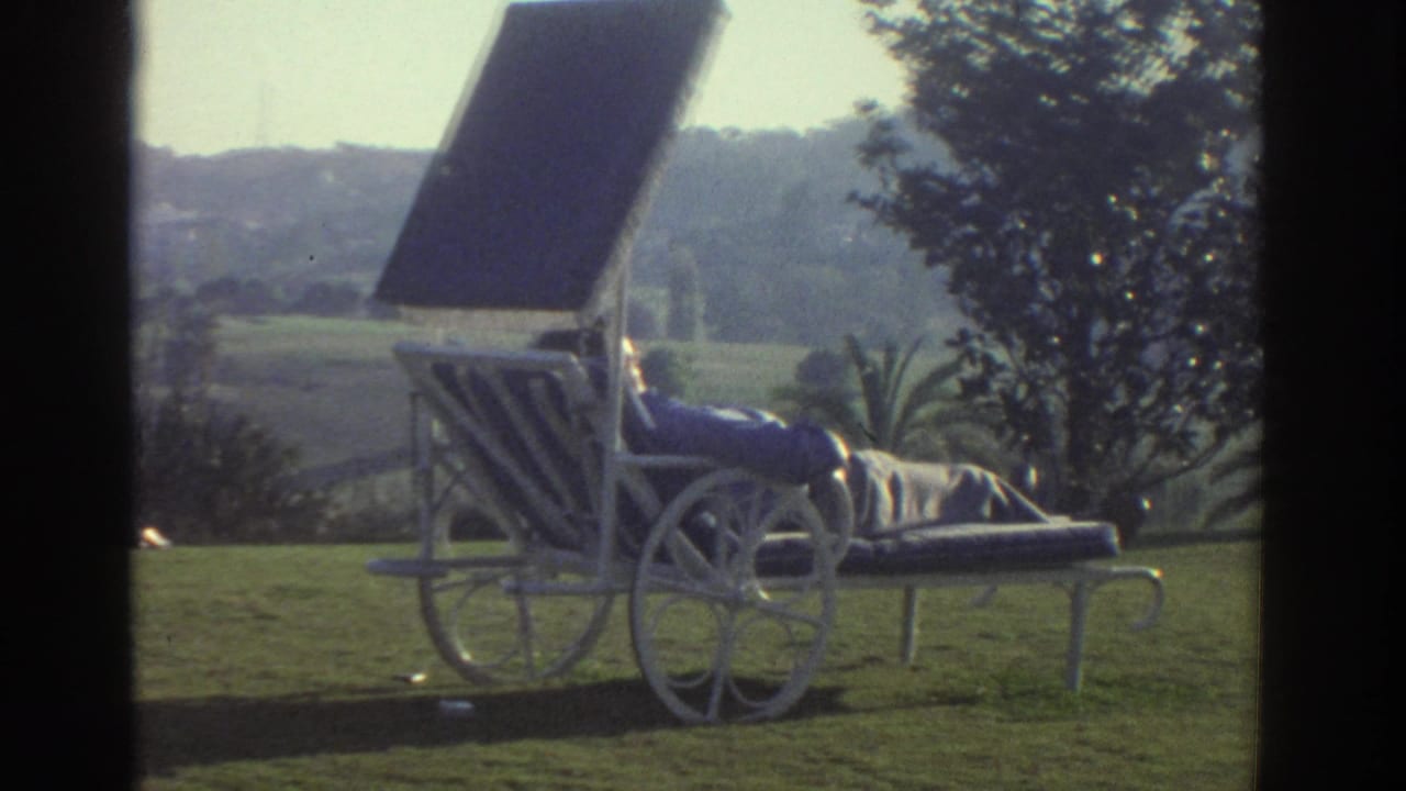 1976 Greece home-movie frame showing a white wheeled lounge chair or cart on grass, with trees, palms, and hills beyond.