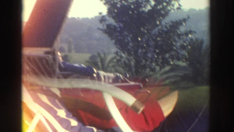 1976 Greece frame showing a person in a red jacket reclining on a white metal chair with a striped cushion beside a pool area.