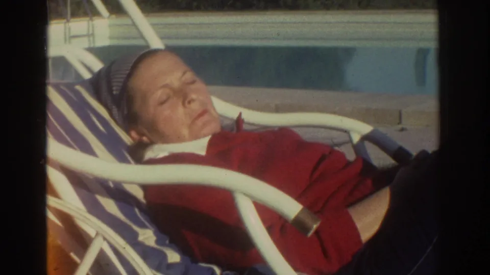 1976 Greece home-movie frame showing a person with eyes closed on a striped lounge chair, with a swimming pool visible behind.