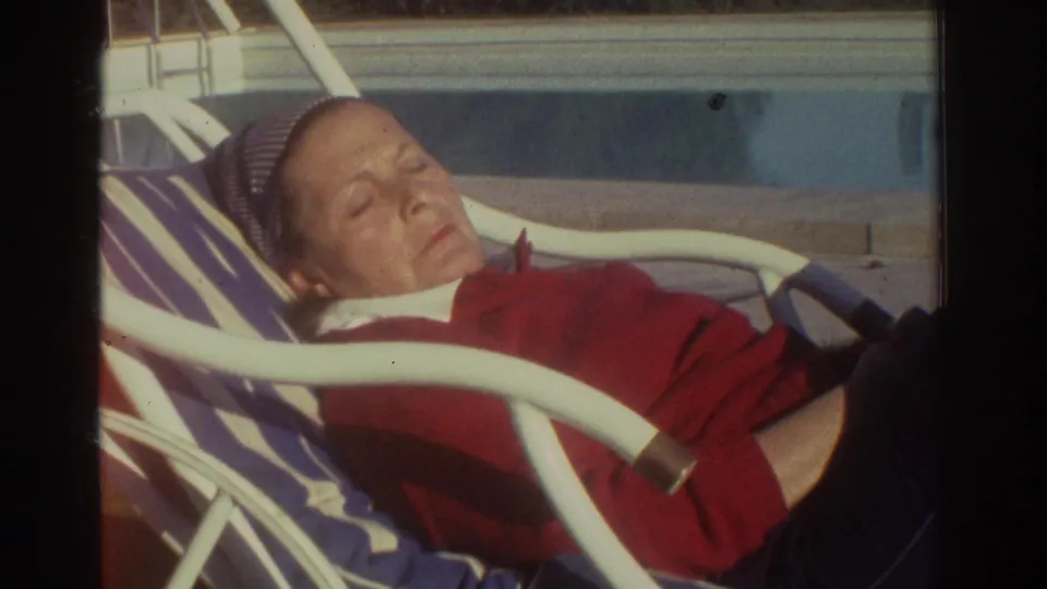1976 Greece frame showing a close poolside view of a person napping on a white metal lounge chair with a striped cushion.