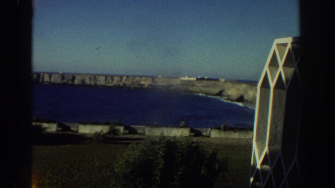 1976 Greece home-movie frame showing blue sea, rocky cliffs, dark window framing, and a white geometric architectural element.