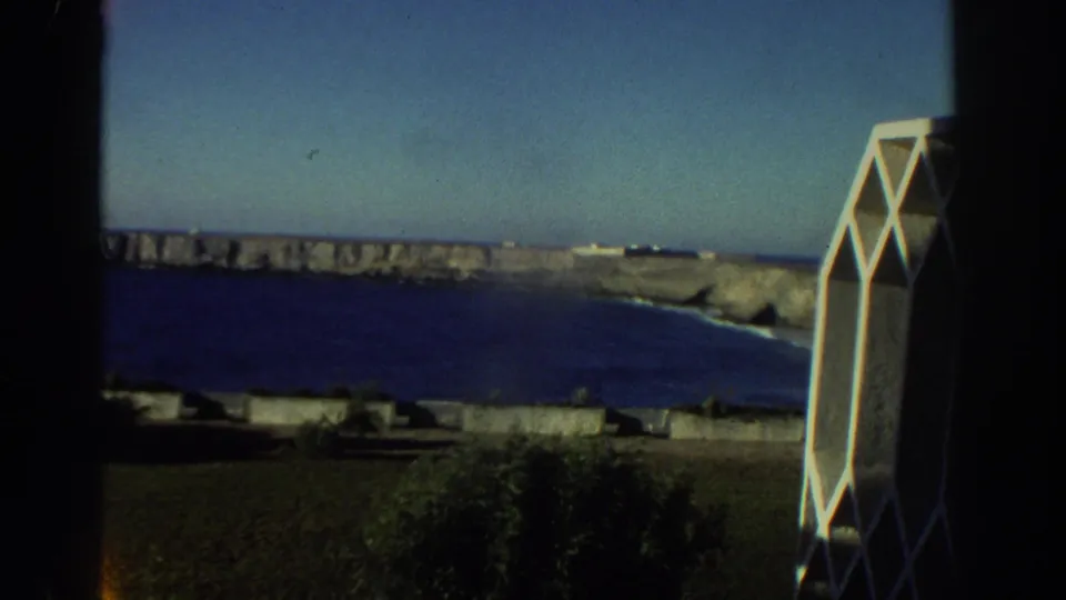 1976 Greece home-movie frame showing blue sea, rocky cliffs, dark window framing, and a white geometric architectural element.