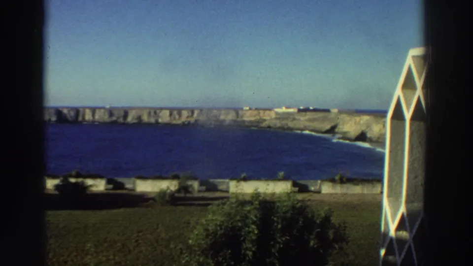A Greece coastal overlook in 1976, with deep blue water, sunlit cliffs, and a white geometric screen near the window edge.