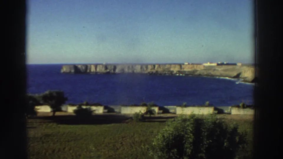 1976 Greece travel footage frames a wide ocean view with pale cliffs, trees, low walls, and open sky from an elevated vantage point.