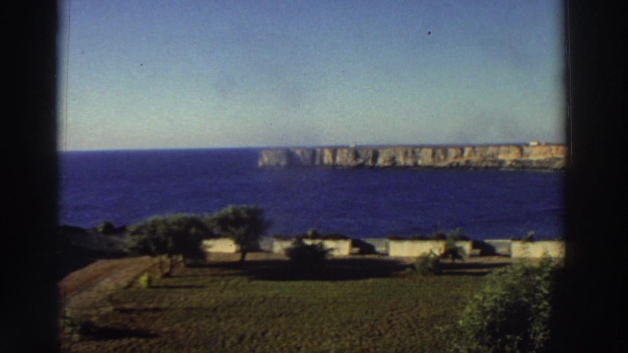 1976 Greece home-movie frame showing a blue sea, distant rocky cliffs, low garden walls, trees, and a dark window divider.