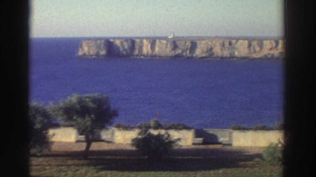 1976 Greece coastal view with a dark vertical window frame, blue water, pale rocky cliffs, low walls, and small trees.