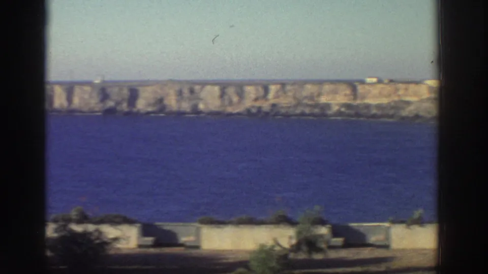 1976 Greece home-movie view across a blue bay toward a long rocky cliff, with low walls and vegetation below.