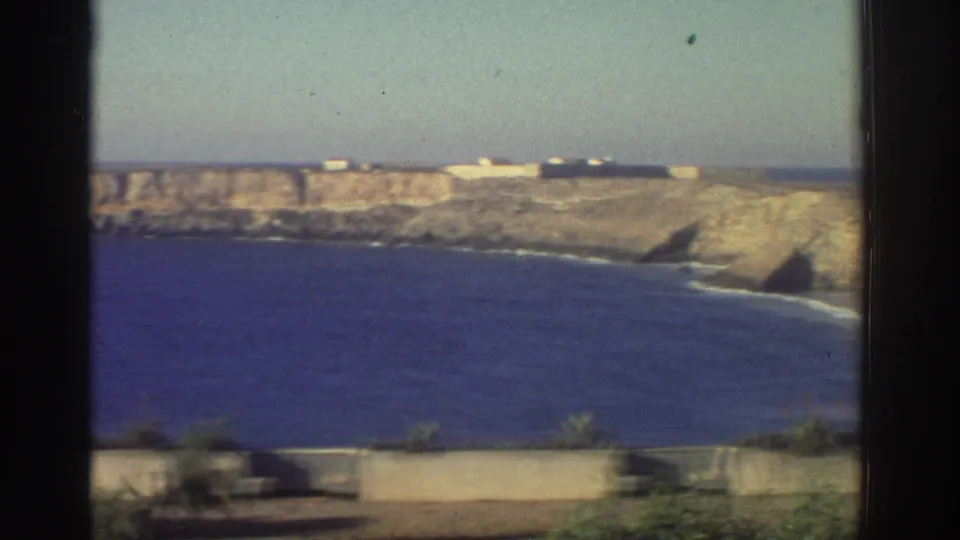1976 Greece coastal frame showing a curving rocky shore, blue water, pale cliff top, low walls, and sparse vegetation.