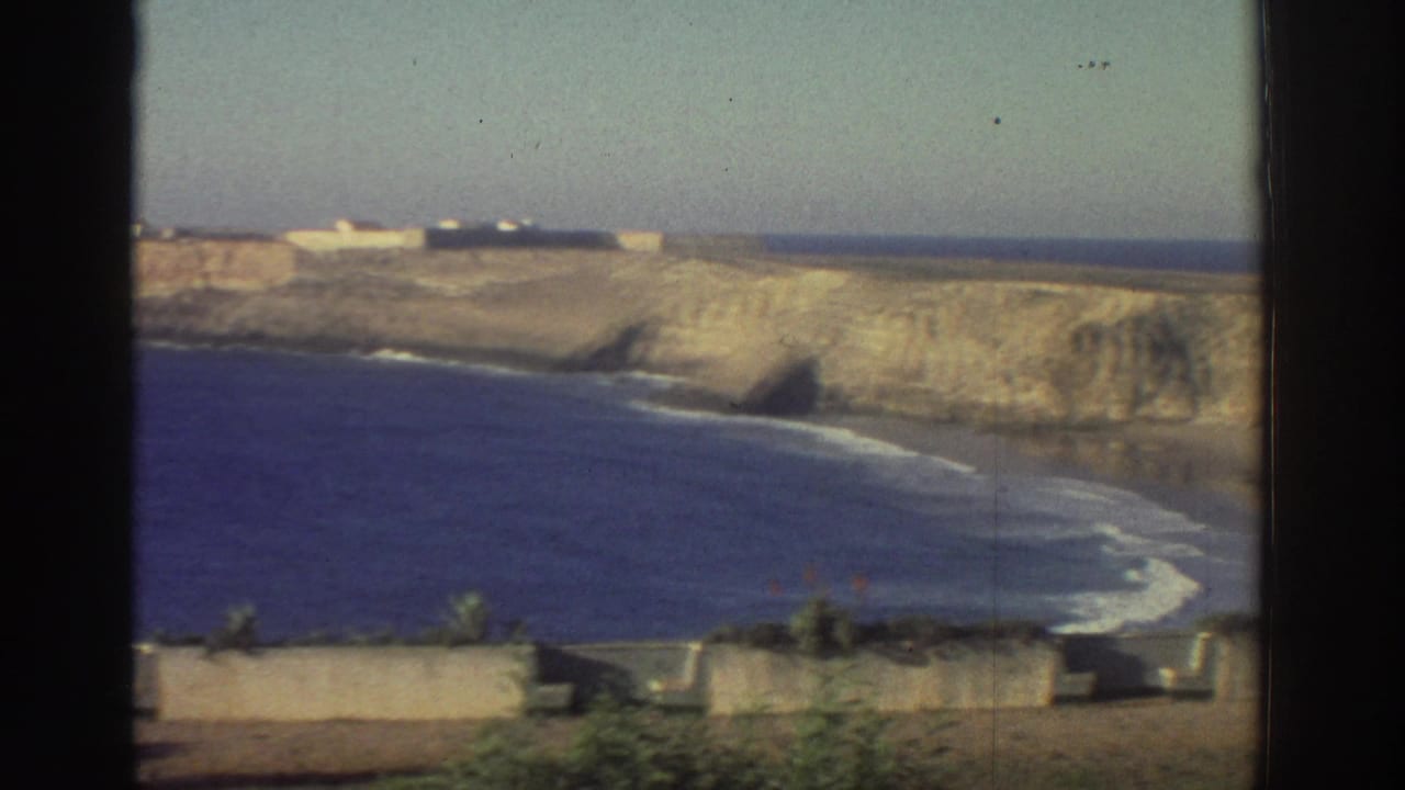 1976 Greece home-movie frame looking past a dark window edge toward coastal cliffs, surf, beach, and a low building on the headland.