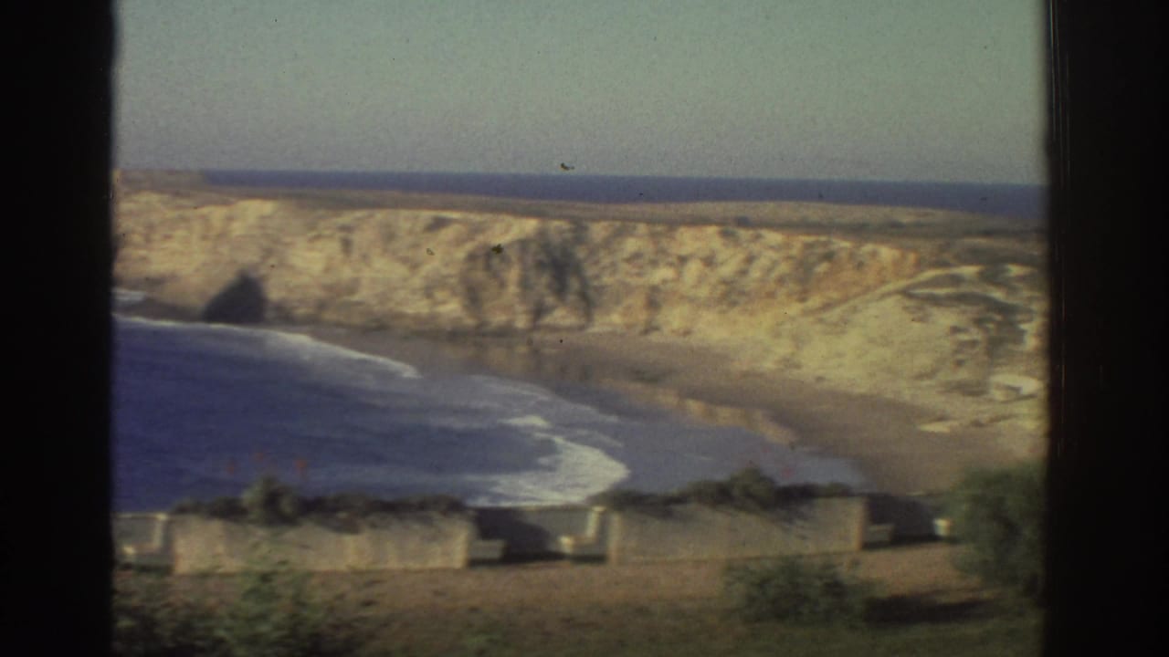 1976 Greece frame showing a sea cove, white surf, pale cliff face, and dark window mullion crossing the view.