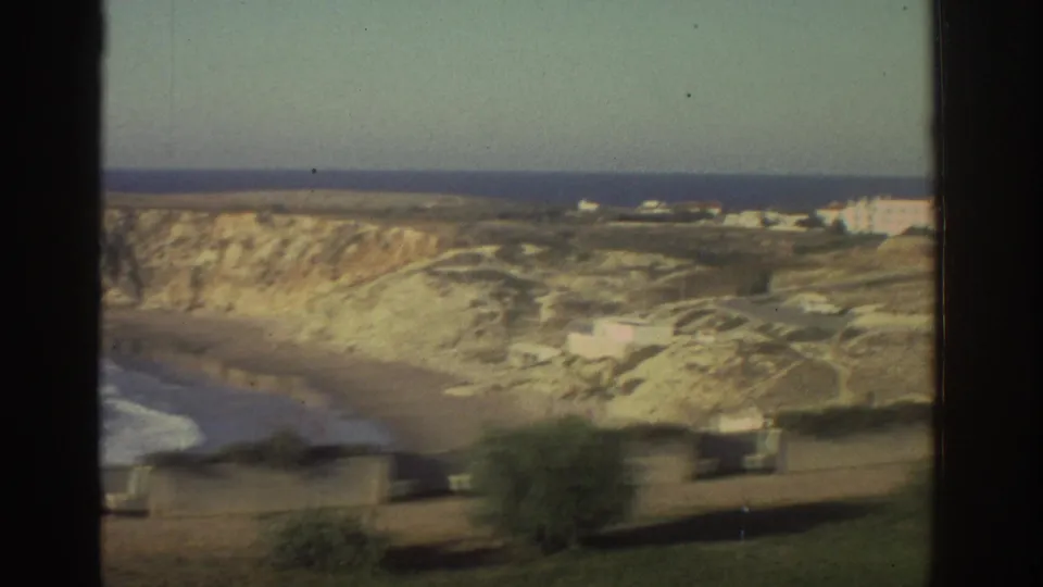 1976 Greece home-movie view of a coastal hillside with beach edge, pale rock, foreground trees, and white buildings near the horizon.