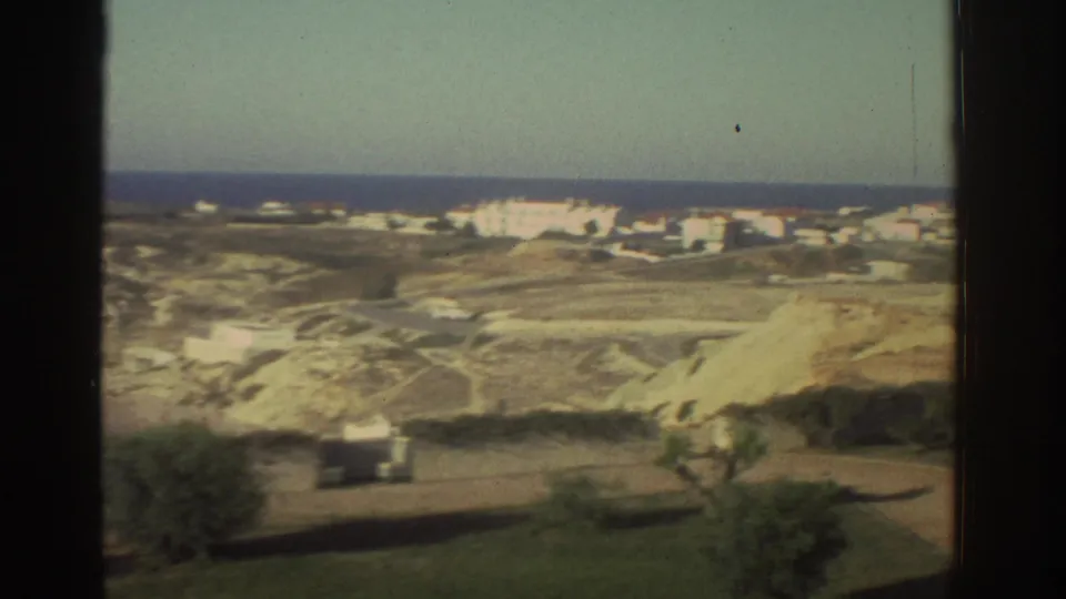 1976 Greece frame of white buildings along a coastal hillside, seen beyond pale cliffs, sparse trees, and the dark window frame.
