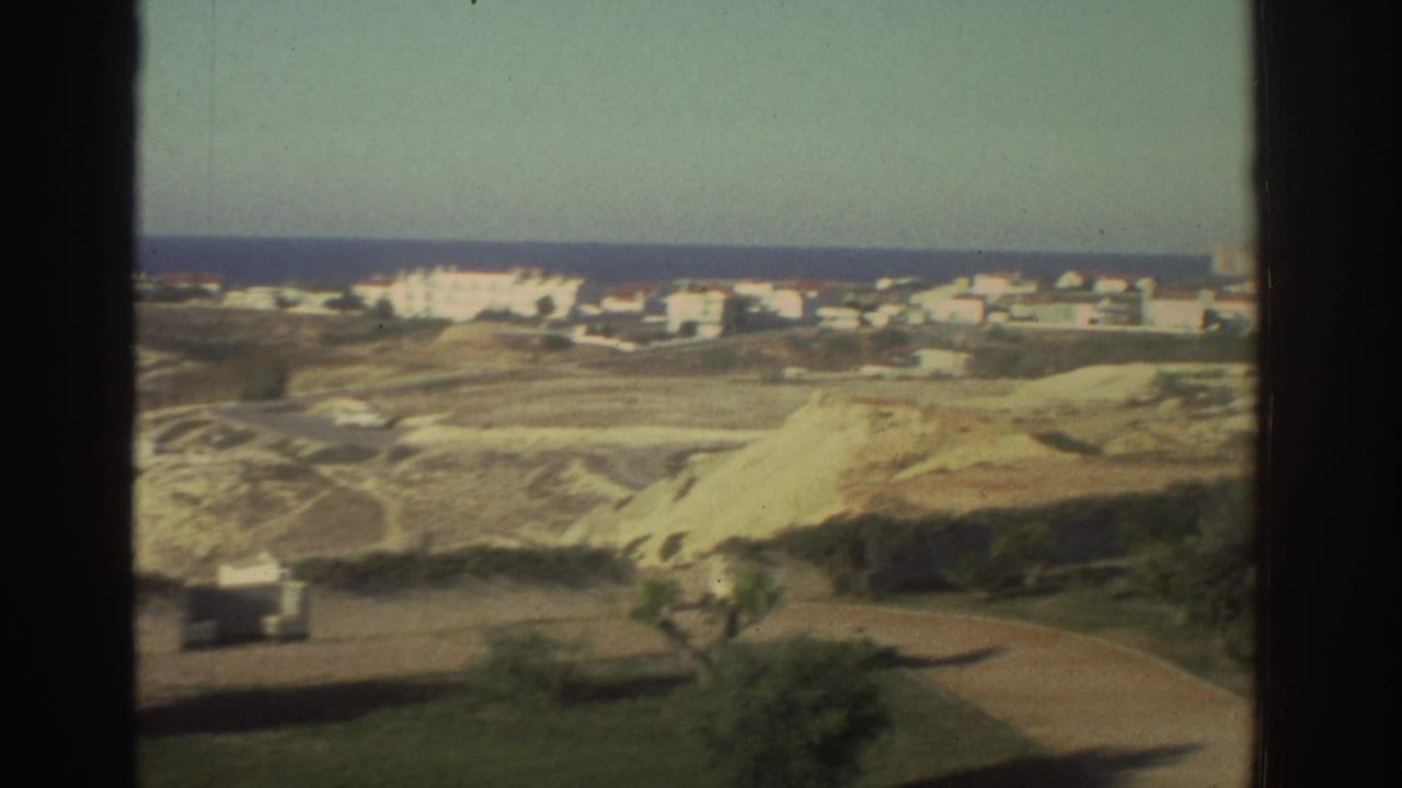 1976 Greece home-movie frame showing a coastal village beyond pale sandy hills, with the sea on the horizon.